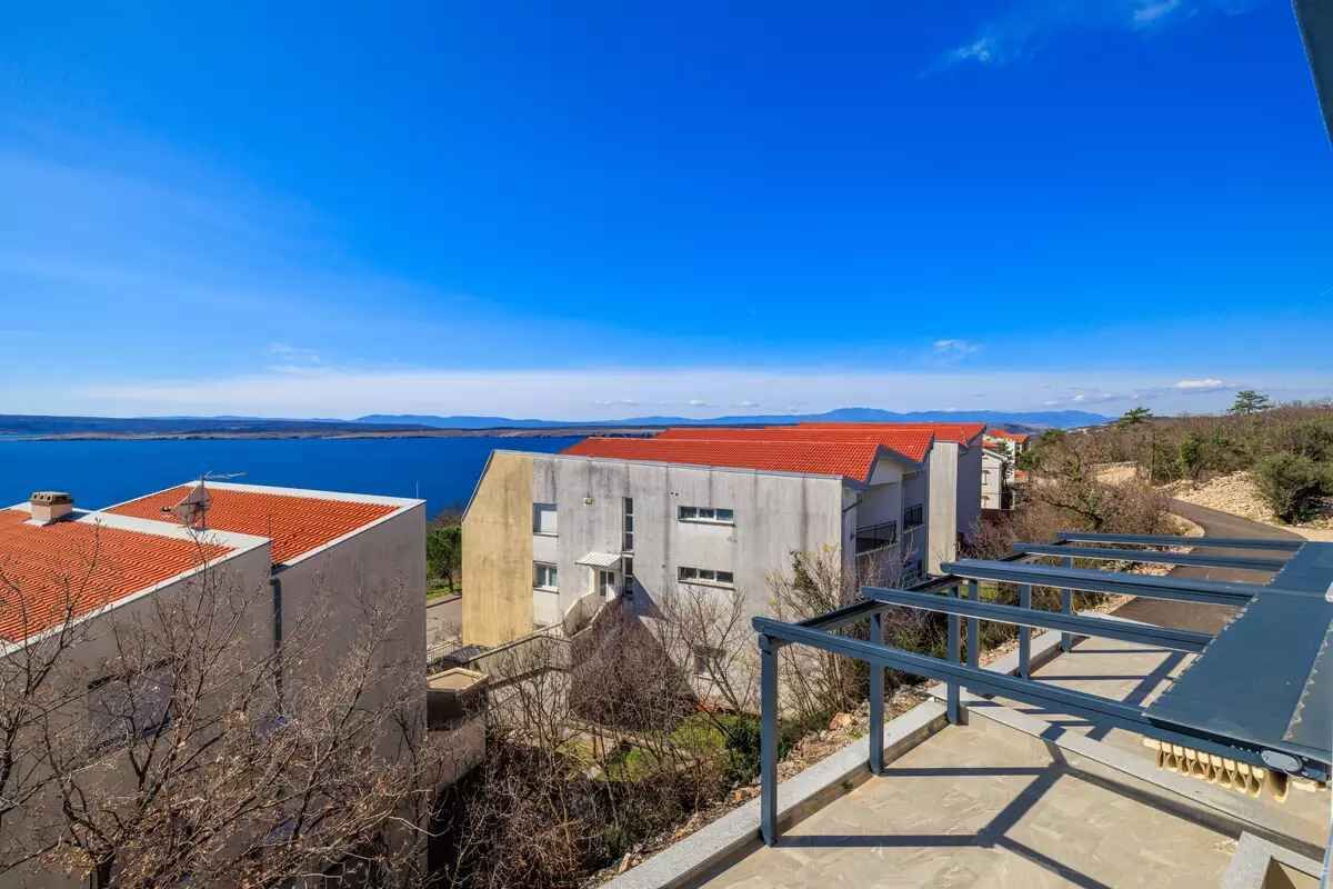 Um edifício costeiro com telhado vermelho, fachada de concreto e vista para o mar e montanhas ao longe.