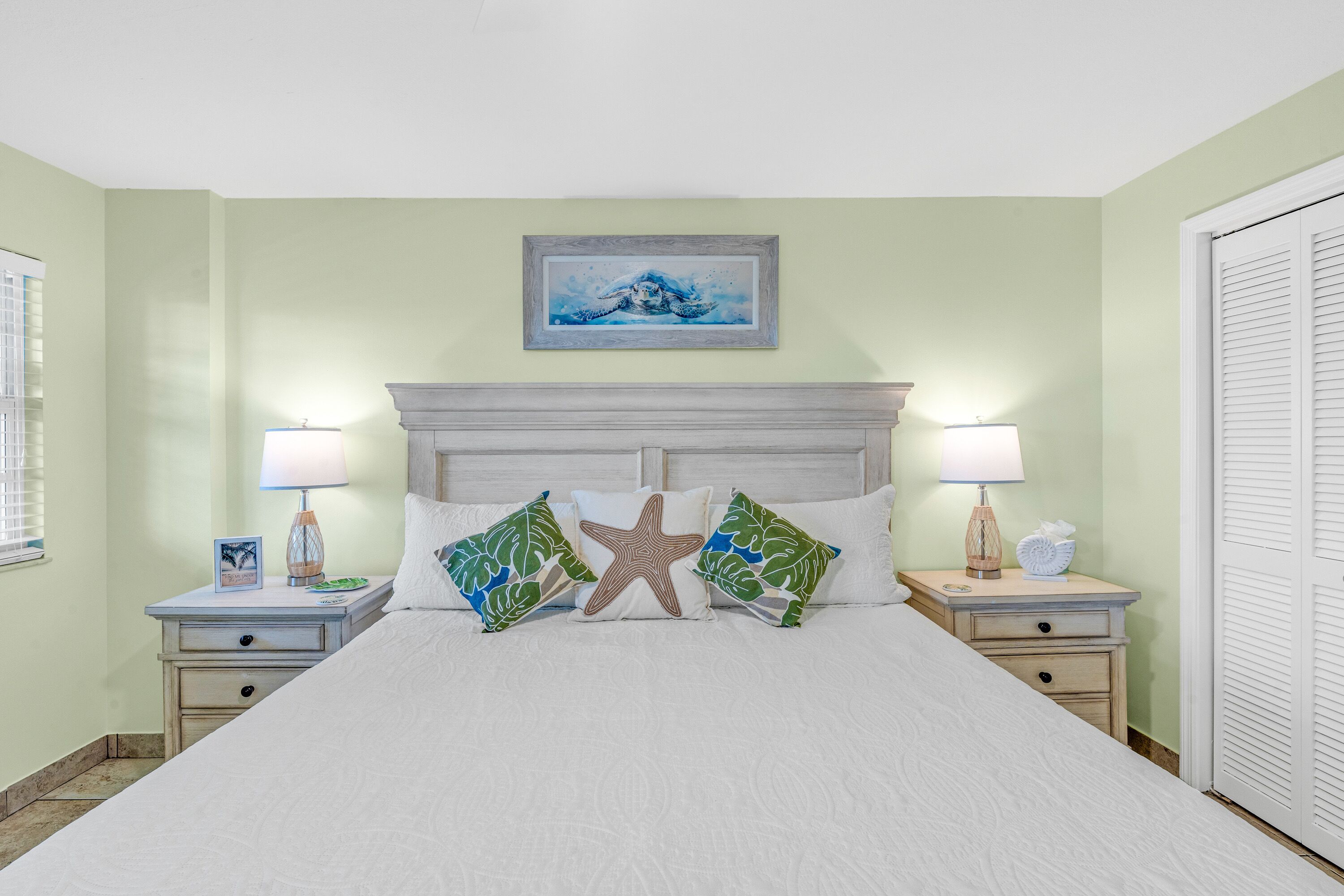 A neatly made bed with a starfish pillow, two nightstands with lamps, a framed picture above the bed, and a closed door on the right.