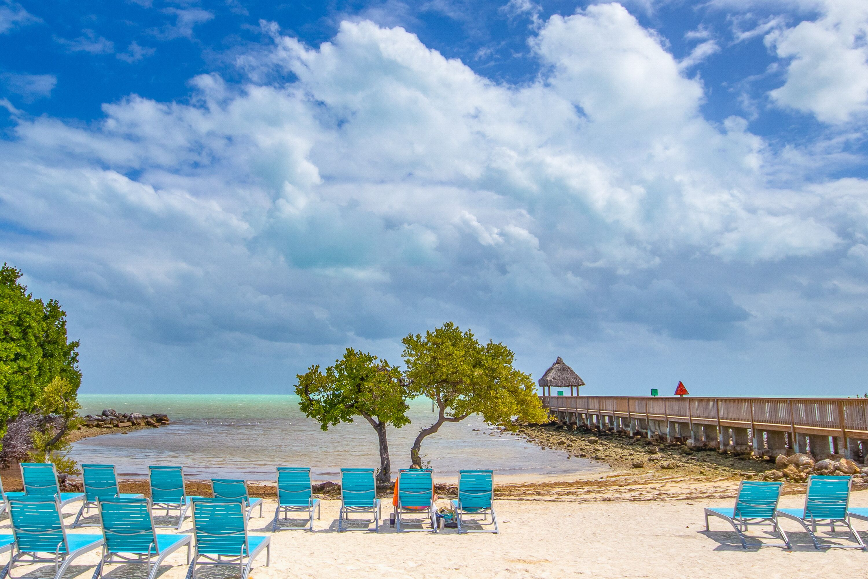 A beach with turquoise chairs, a wooden pier, and a thatched-roof structure.