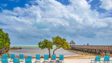 A beach with turquoise chairs, a wooden pier, and a thatched-roof structure.
