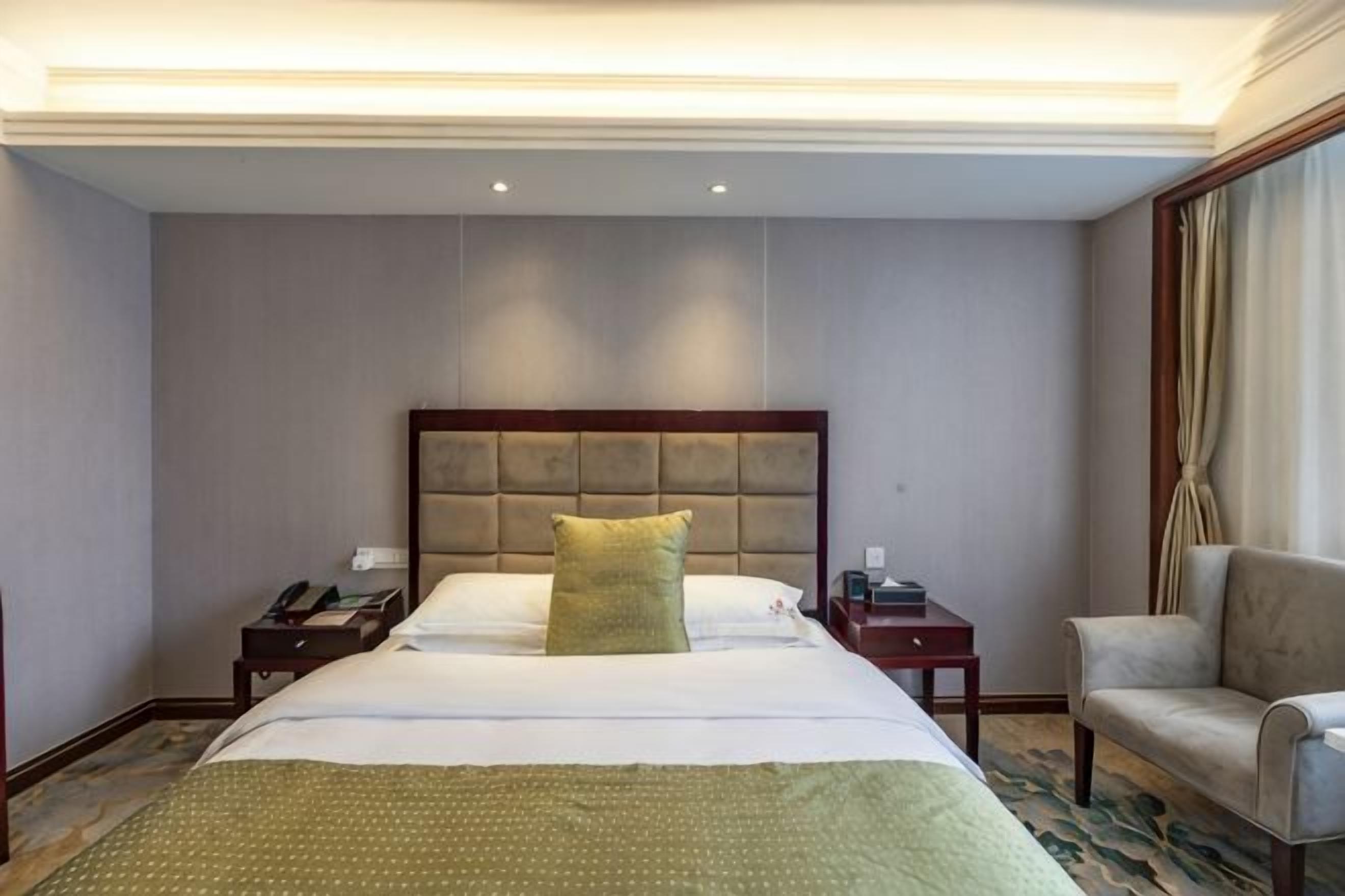 A hotel room with a large bed, a bedside table with a lamp, a chair, and a window with curtains.