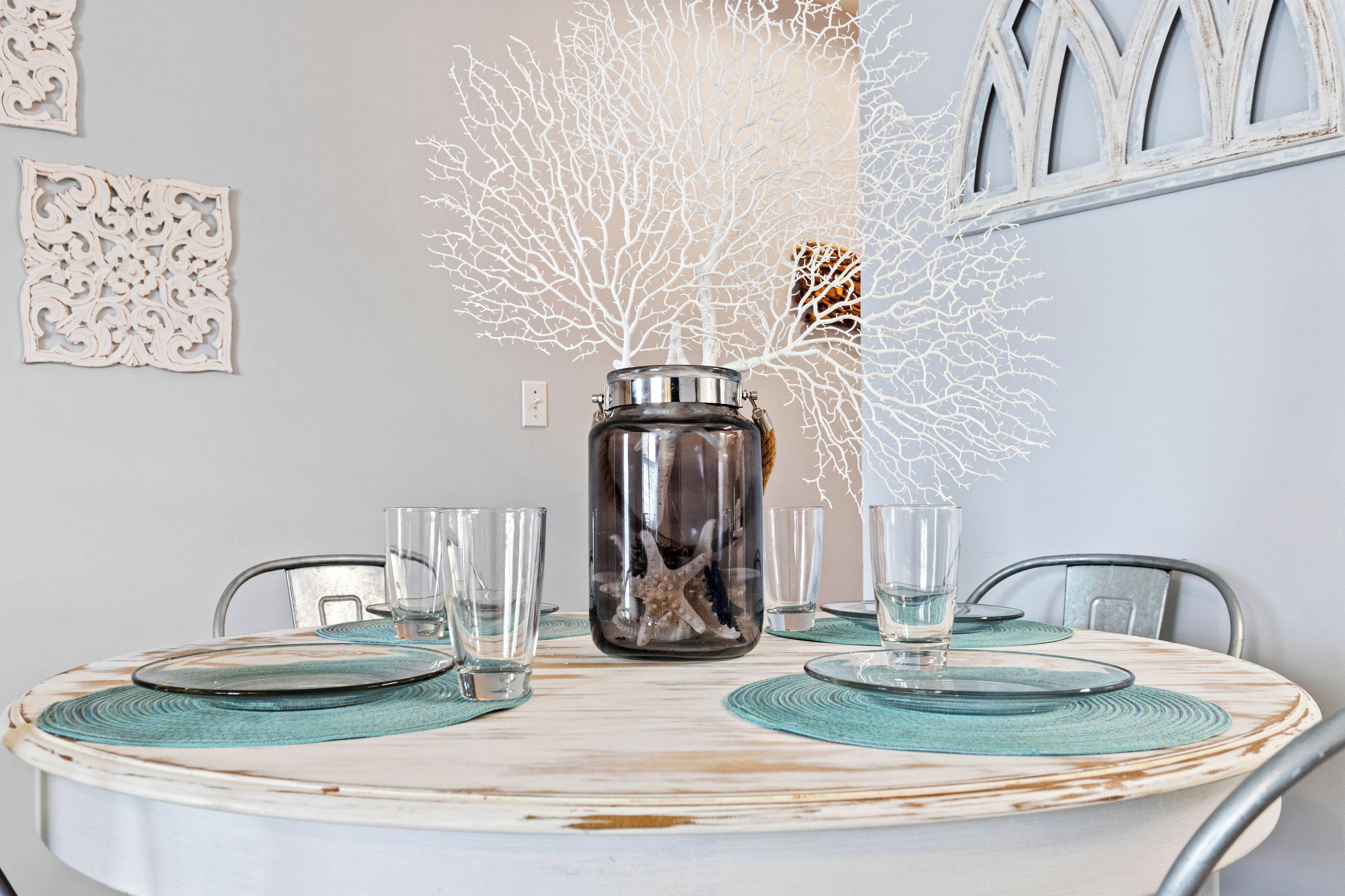 A round dining table with a glass vase containing a starfish and coral, surrounded by clear glasses and teal placemats.