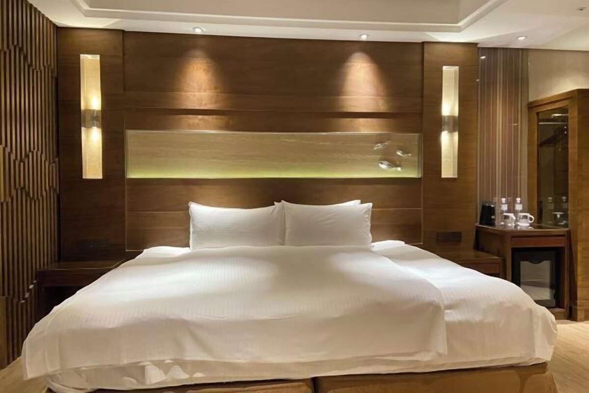 Quarto de hotel moderno com uma cama grande, painéis de madeira e iluminação embutida.
