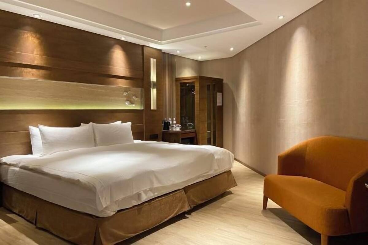 A modern hotel room with a large bed, a wooden headboard, a wooden floor, and a single orange armchair.