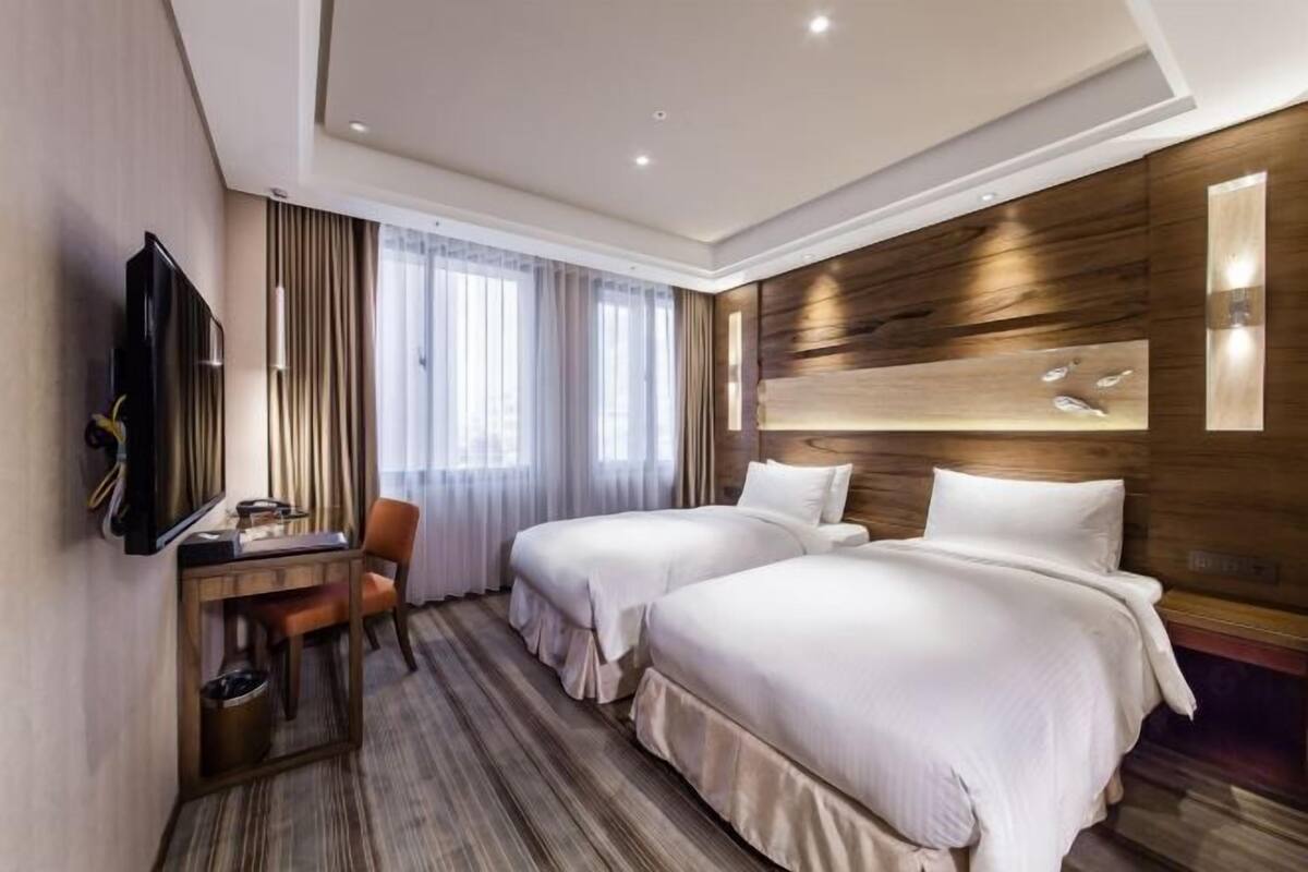 A hotel room with two beds, a desk, a chair, a TV, and a large window with curtains.