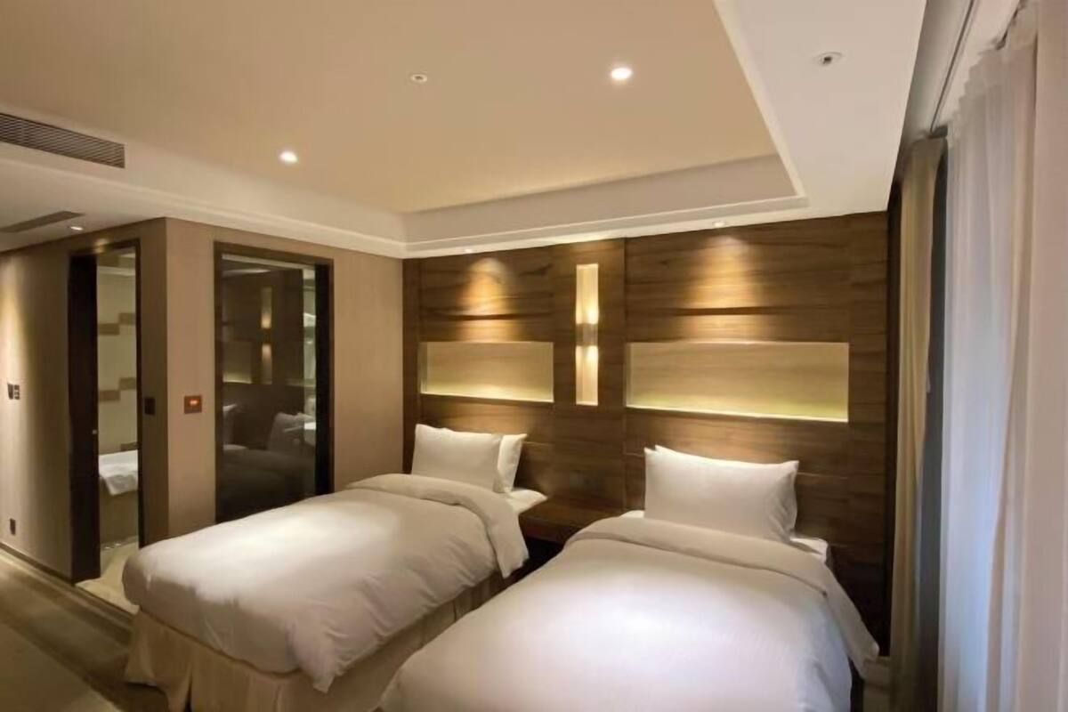 A hotel room with two beds, a wooden accent wall, and a glass door leading to a bathroom.