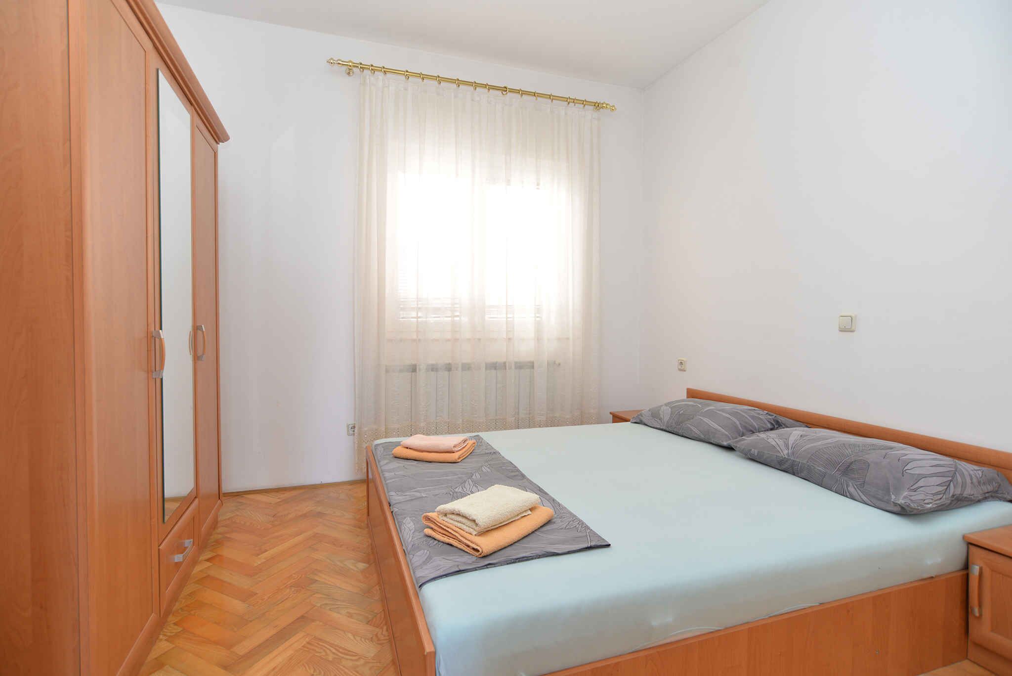 A bedroom with a bed, a wooden wardrobe, a window with sheer curtains, and a wooden floor.