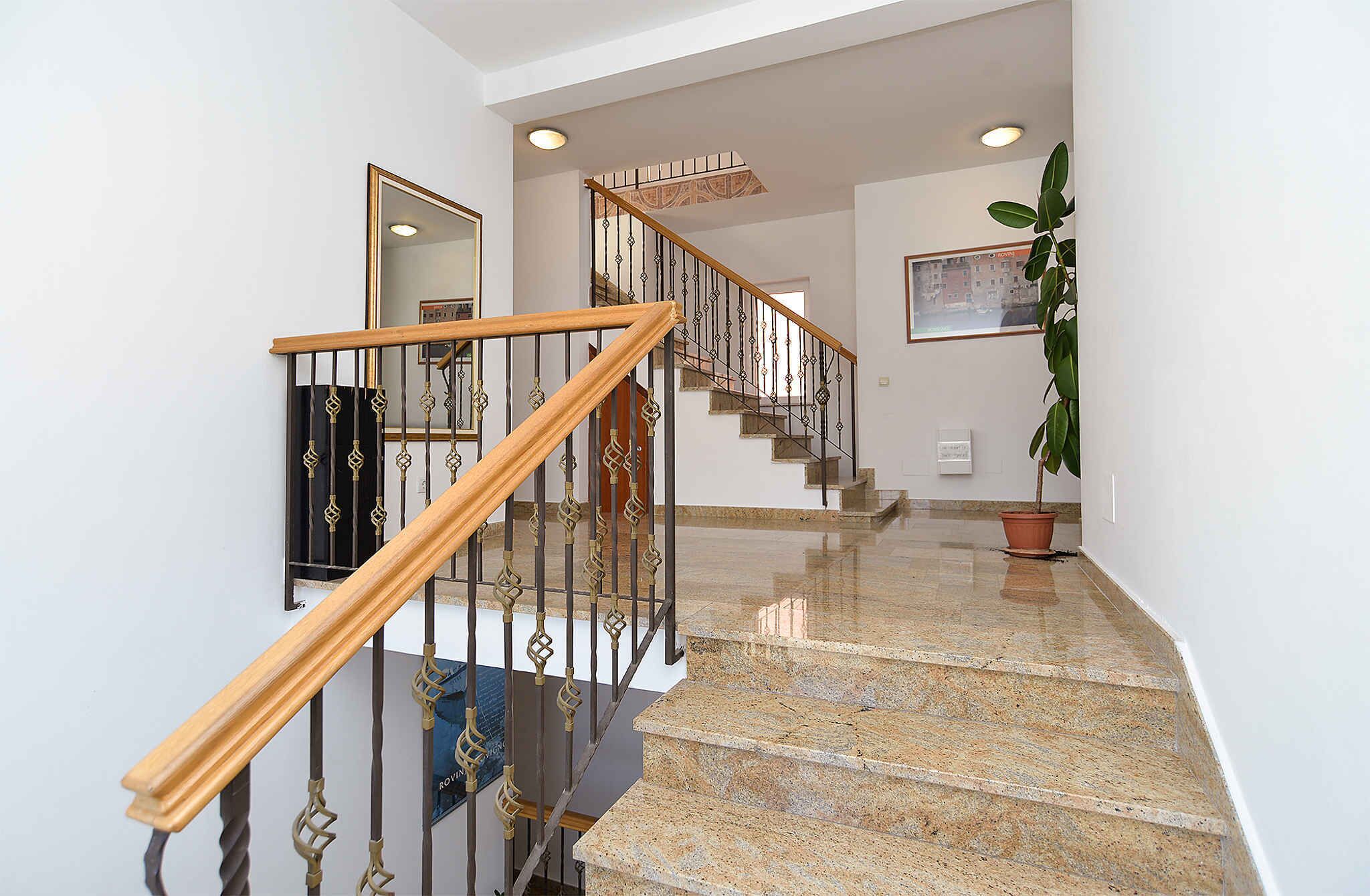 A polished granite staircase with a wooden handrail and metal balusters, leading to a landing with a framed picture and a potted plant.