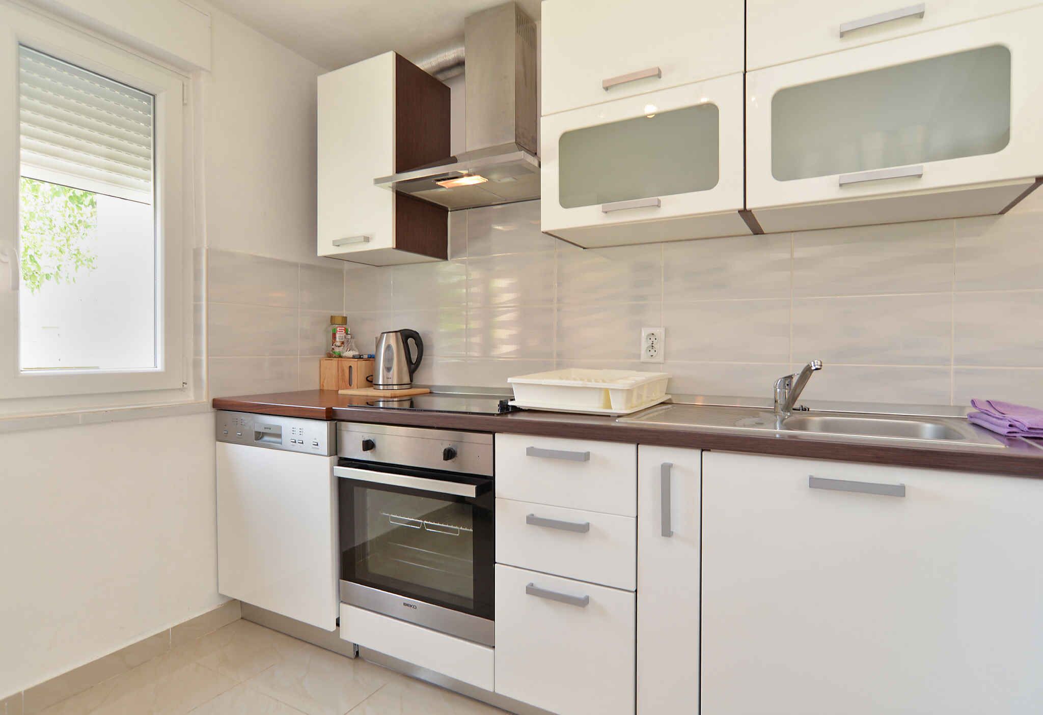 A modern kitchen with white cabinets, a stainless steel oven, a dishwasher, and a window with blinds.