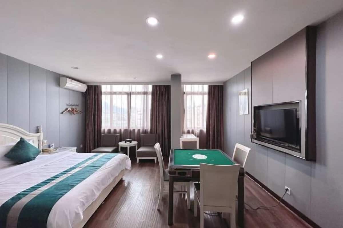 A hotel room with a bed, a desk with a green table, a TV mounted on the wall, and a window with curtains.