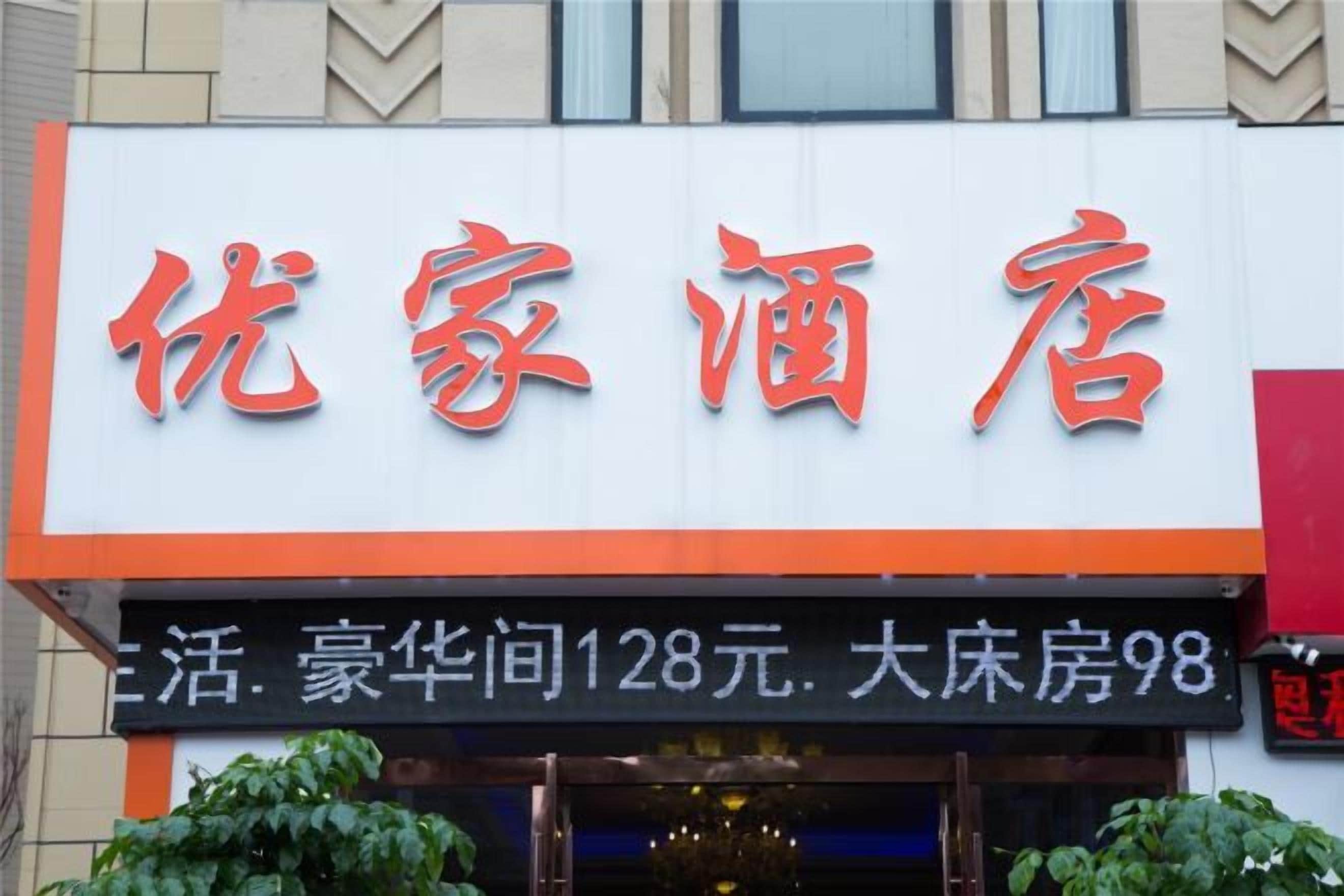 Letrero de hotel con el nombre '优家酒店' en grandes caracteres rojos sobre un fondo blanco con un borde naranja. Debajo del letrero, hay otro con texto blanco que indica las tarifas de las habitaciones.