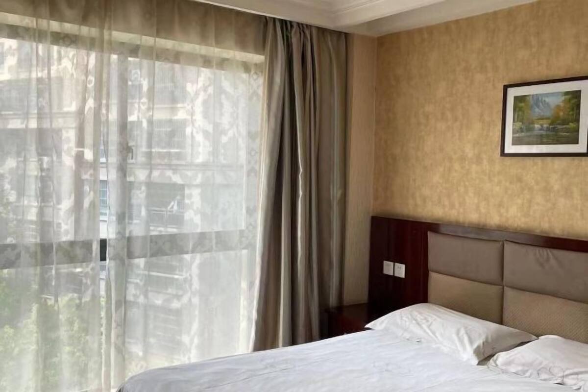 A hotel room with a bed, curtains, a painting on the wall, and a view of trees through the window.