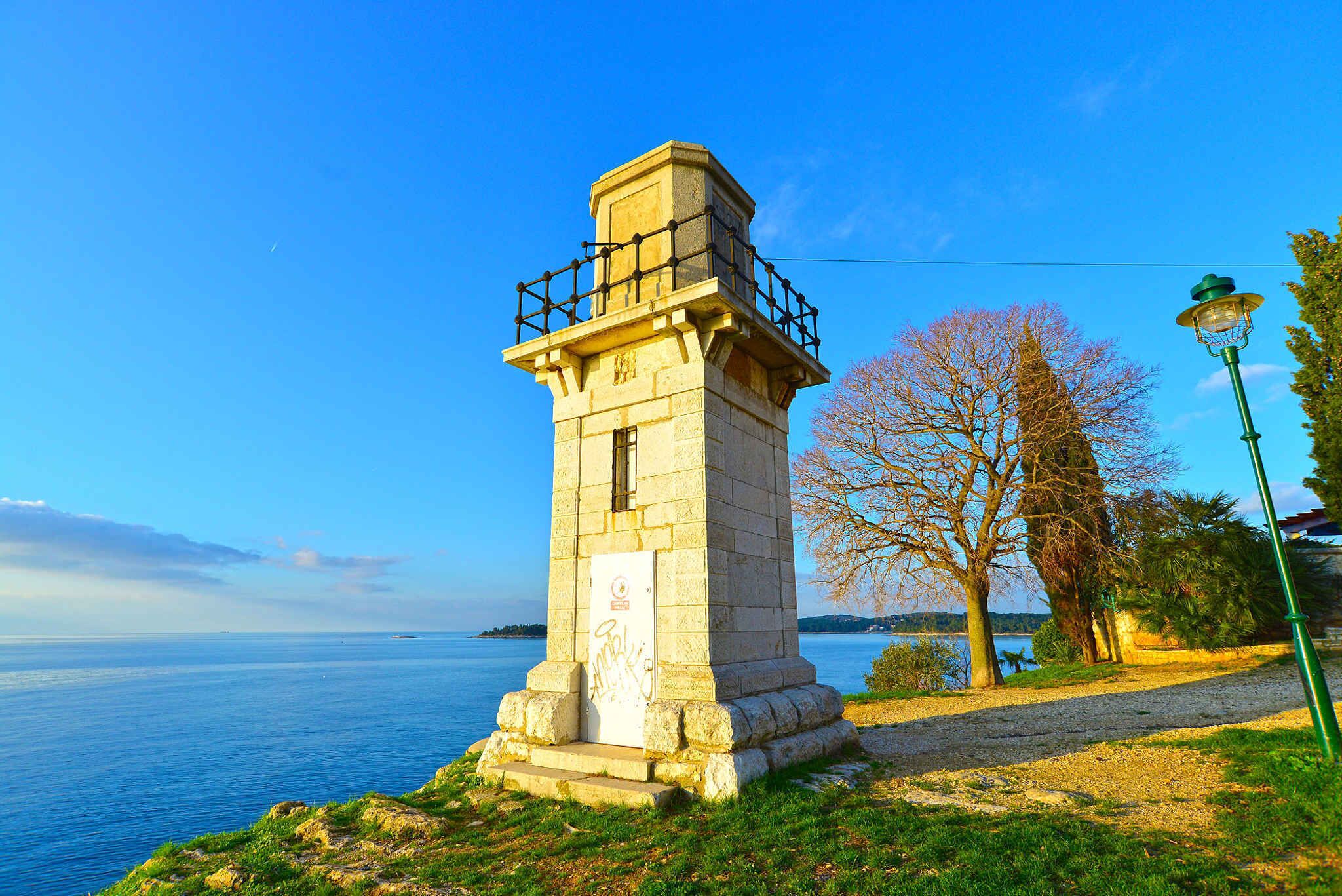 A stone lighthouse with a metal railing on top, situated by a body of water with a clear sky.