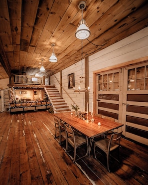 Un interior rústico de madera con una larga mesa de comedor, sillas de madera y lámparas colgantes. Una escalera conduce a un nivel superior con un balcón.