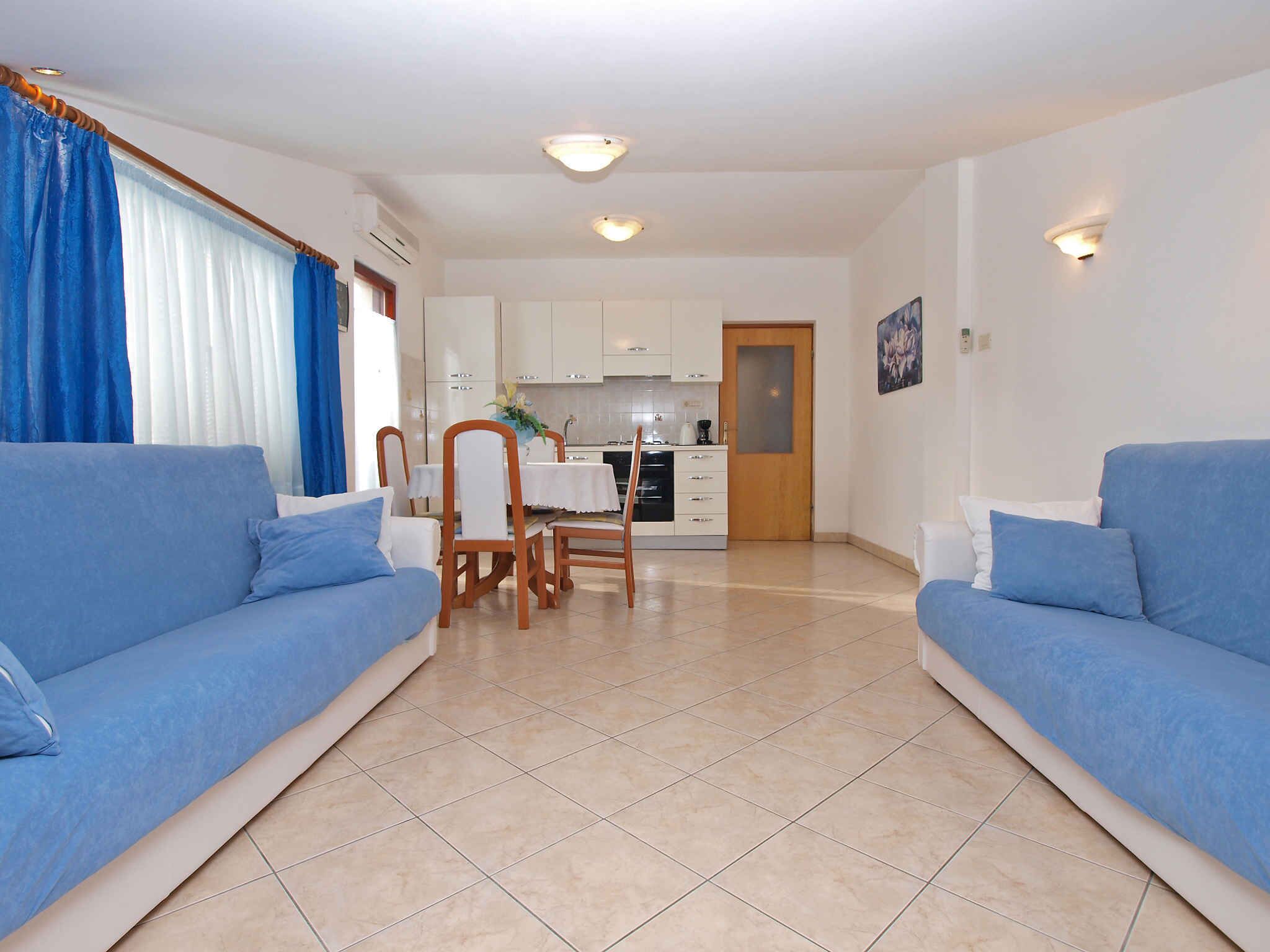 A living room with two blue sofas, a dining table with chairs, a kitchen area, and a door leading to another room.