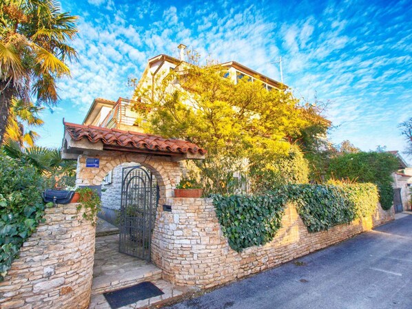 A stone-walled building with a tiled roof, a gated entrance, and lush greenery. - Holiday apartment with air conditioning (Medulin)