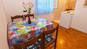 A small dining area with a table covered in a colorful balloon pattern, wooden chairs, and a vase with flowers. A kitchen is visible in the background with a refrigerator and a sink.