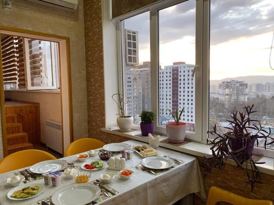 Un comedor con una mesa dispuesta para cuatro personas, plantas en macetas en el alféizar de la ventana y vistas de un paisaje urbano a través de amplios ventanales.