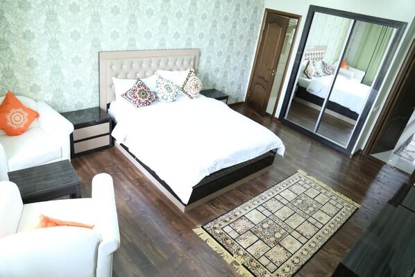 A bedroom with a large bed, a nightstand, a chair, a mirror, and a patterned rug. - Sunrise Apartment (Dushanbe)