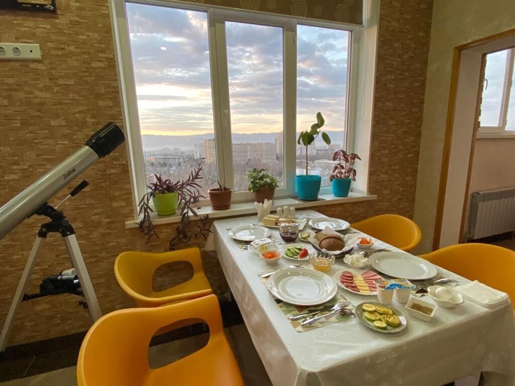 A dining table set with plates, cutlery, and food, with a telescope and potted plants on a balcony with a city view.