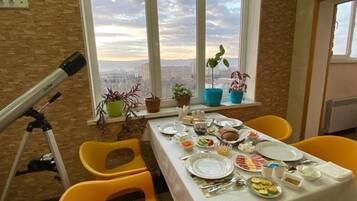 A dining table set with plates, cutlery, and food, with a telescope and potted plants on a balcony with a city view.