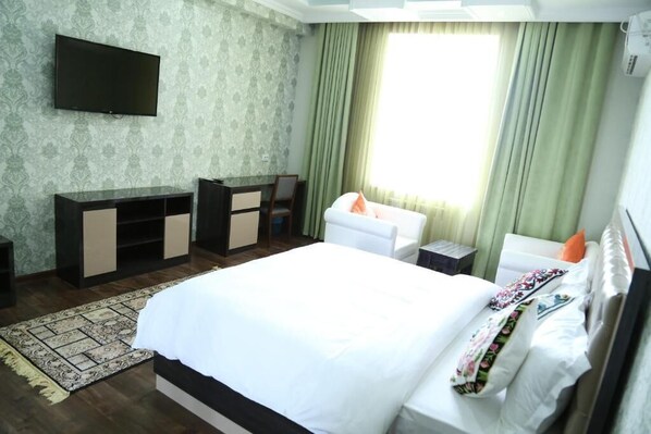 A hotel room with a large bed, a TV mounted on the wall, a desk with a chair, and a window with curtains. - Sunrise Apartment (Dushanbe)