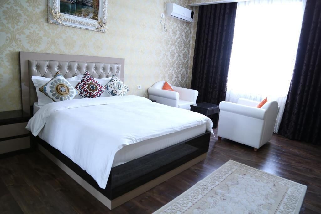 Un dormitorio con una cama grande, dos sillones blancos, una mesita de noche oscura y un ventanal con cortinas.