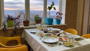 Tavola imbandita per la colazione con diversi piatti, piante in vaso e vista sulla città attraverso ampie finestre.