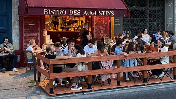 Un espacio al aire libre concurrido del BISTRO DES AUGUSTINS, con un toldo rojo y un cartel de menú visible.