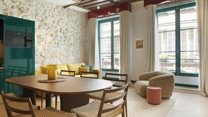 Un comedor con una mesa redonda, sillas y un sofá amarillo. Se aprecia una pared de piedra detrás del sofá y una ventana con cortinas.