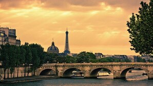 Un puente de piedra con múltiples arcos sobre un río, con la Torre Eiffel y una cúpula al fondo.