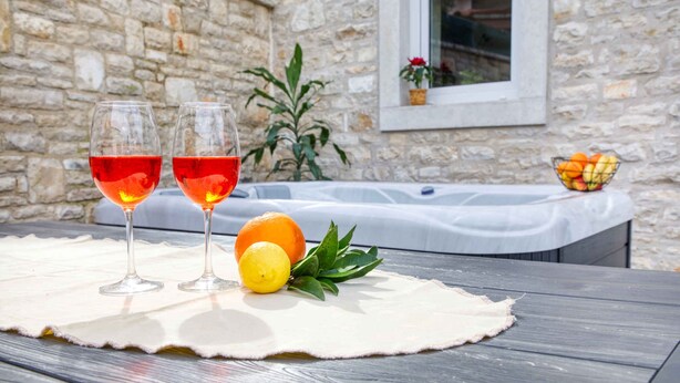 Two glasses of red wine on a table with a placemat, a lemon, and an orange. A hot tub is visible in the background. - Holiday home with air conditioning (Peroj)