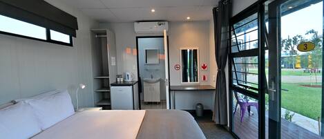 A hotel room with a bed, a desk with a chair, a small kitchenette, a bathroom, and a large window with a view of the outside.