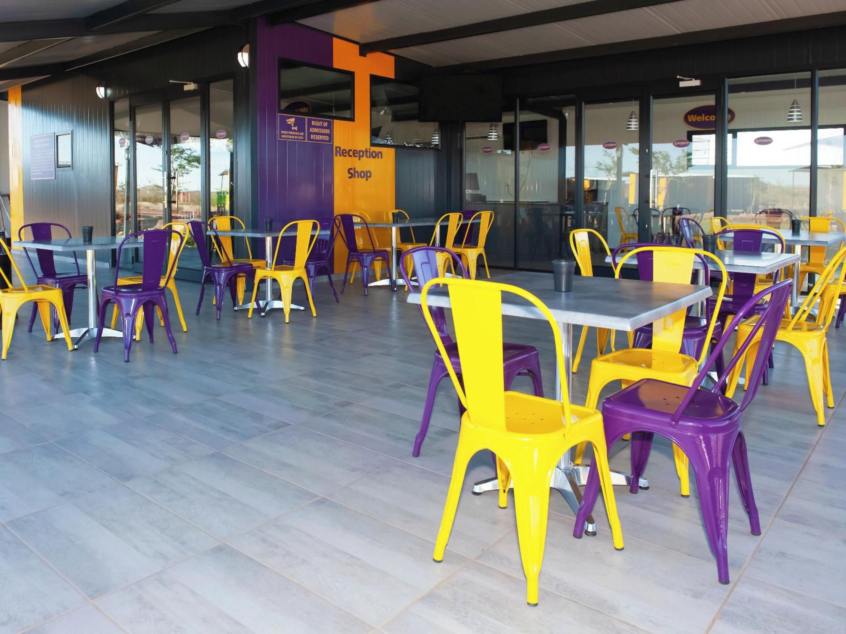 Uma área de recepção com cadeiras e mesas em roxo e amarelo, um balcão de recepção e um aviso indicando que o acesso é reservado.