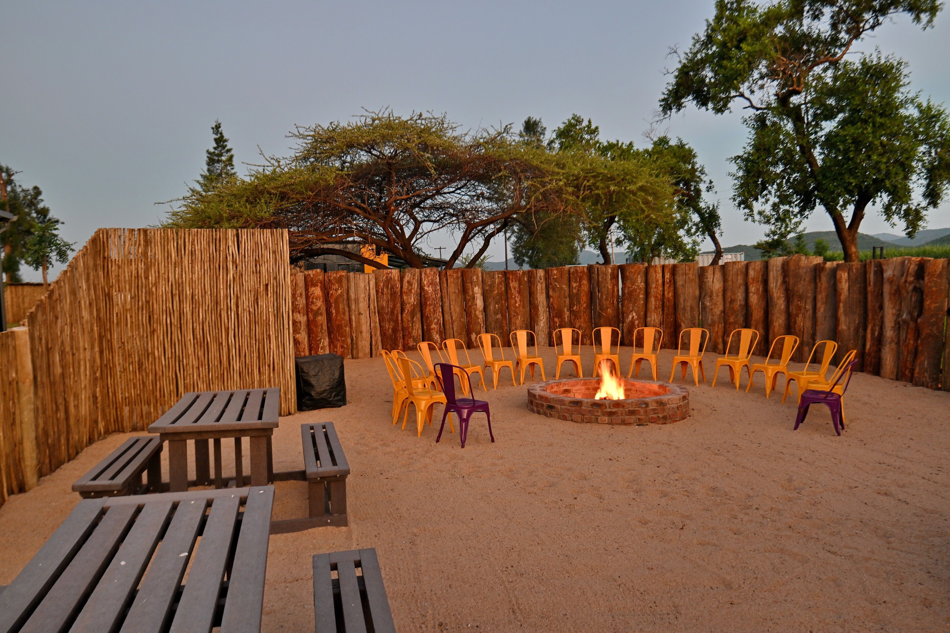 An outdoor seating area with wooden benches and plastic chairs arranged around a central fire pit.