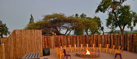An outdoor seating area with wooden benches and plastic chairs arranged around a central fire pit.