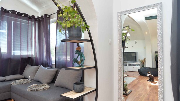 Een moderne woonkamer met een grijze hoekbank, een metalen plank met een plant en een grote spiegel die de kamer weerkaatst.