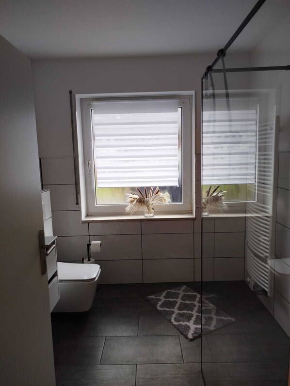 Un baño con inodoro, una ventana con persianas, zona de ducha y un tapete estampado en el suelo.