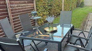 An outdoor patio with a glass-top table, four chairs, and a small potted plant. - Modern and family-friendly: 3 bedrooms in Burhave (6 pers. plus baby crib) (Butjadingen)