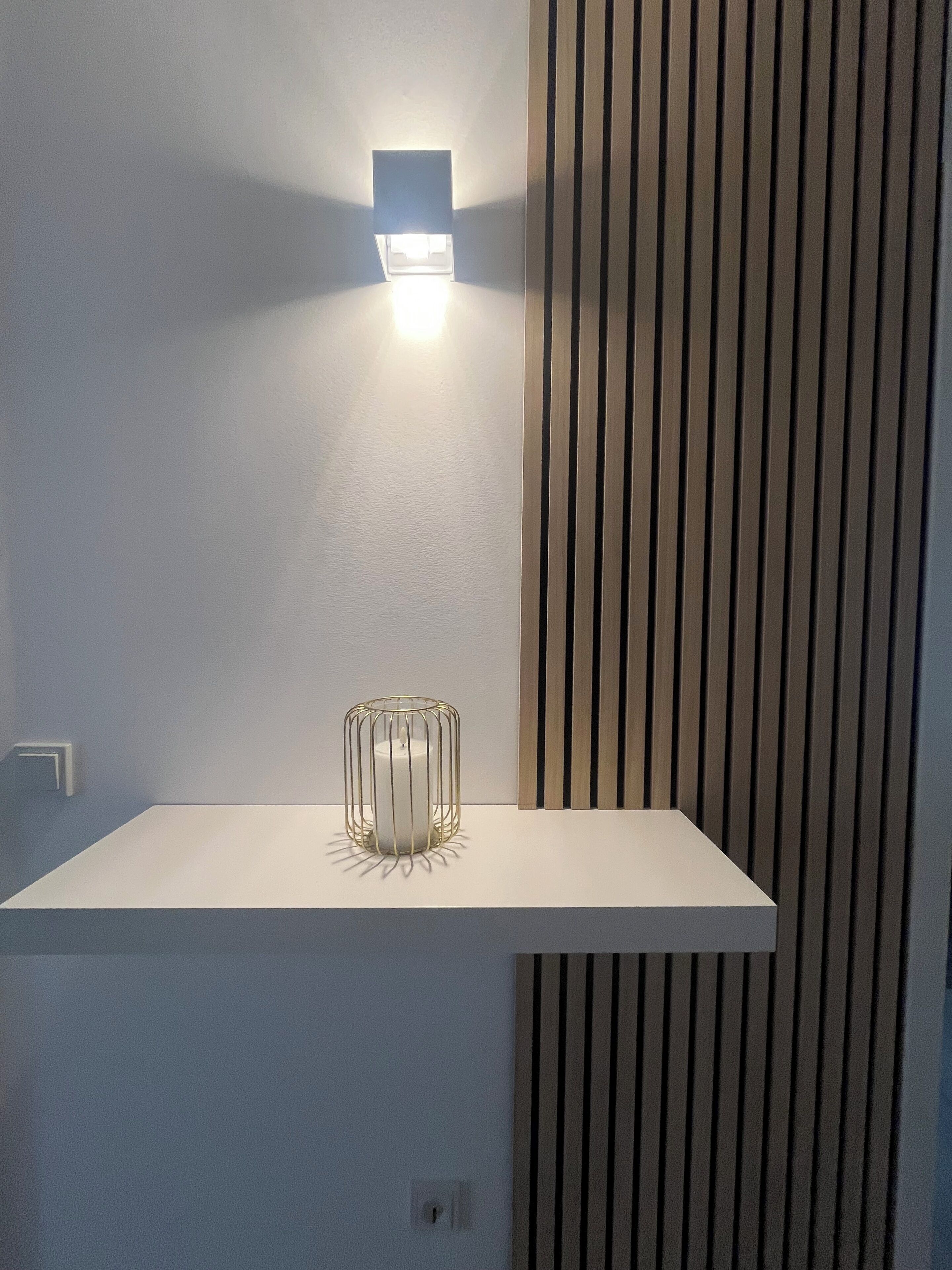 A white shelf with a candle in a wire cage, a wall-mounted light, and a vertical wooden panel.