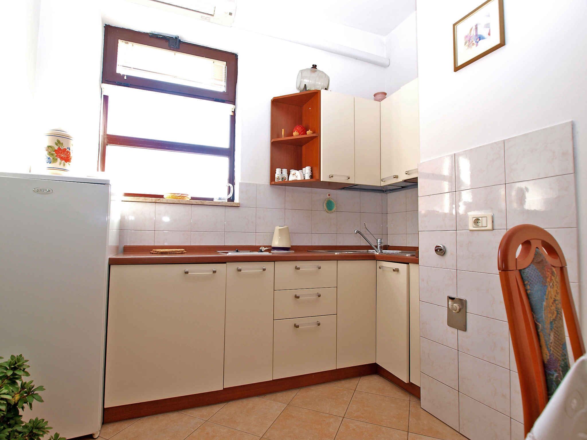 A compact kitchen with white cabinets, a wooden countertop, a window with blinds, and a small shelf with kitchen items.