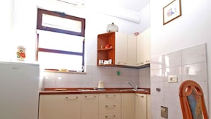 Een kleine keuken met witte kastjes, een houten aanrechtblad, een raam met jaloezieën en een klein plankje met keukenspullen.