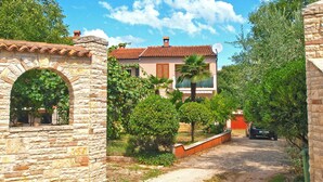 A stone archway leading to a house with a tiled roof, surrounded by greenery and a gravel path.