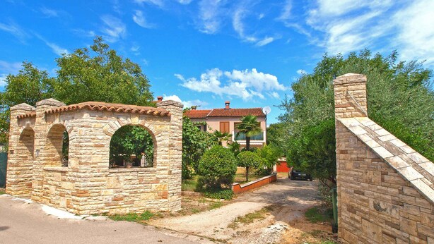 A stone structure with arches, a paved road, and a building with a tiled roof in the background.