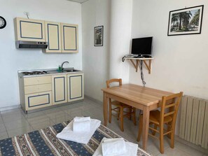 A compact kitchen with a stove, sink, and cabinets, a dining table with chairs, and a TV on a stand.