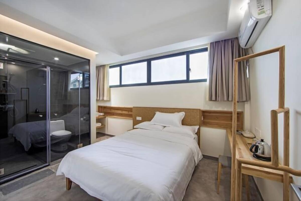 Ein Hotelzimmer mit Bett, Nachttisch und einer Glasscheibe, die den Raum vom Badezimmer trennt.