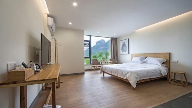 A modern hotel room with a wooden floor, a bed with white bedding, a desk with a chair, a small table, and a large window with a mountain view. - Wuyishan Yunzhong Jinshu Homestay (Wuyishan)