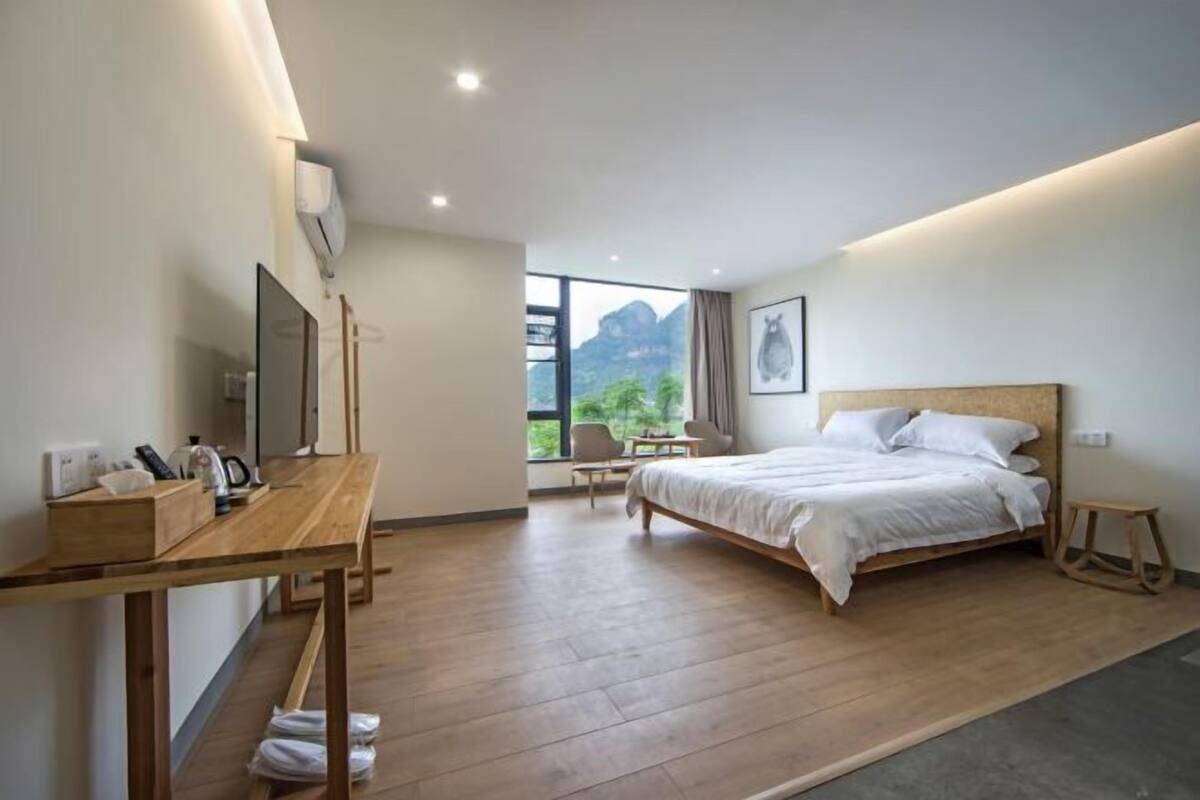 Ein modernes Hotelzimmer mit Holzboden, einem Bett mit weißer Bettwäsche, einem Schreibtisch mit Stuhl, einem kleinen Tisch und einem Fenster mit Blick auf Grünflächen.