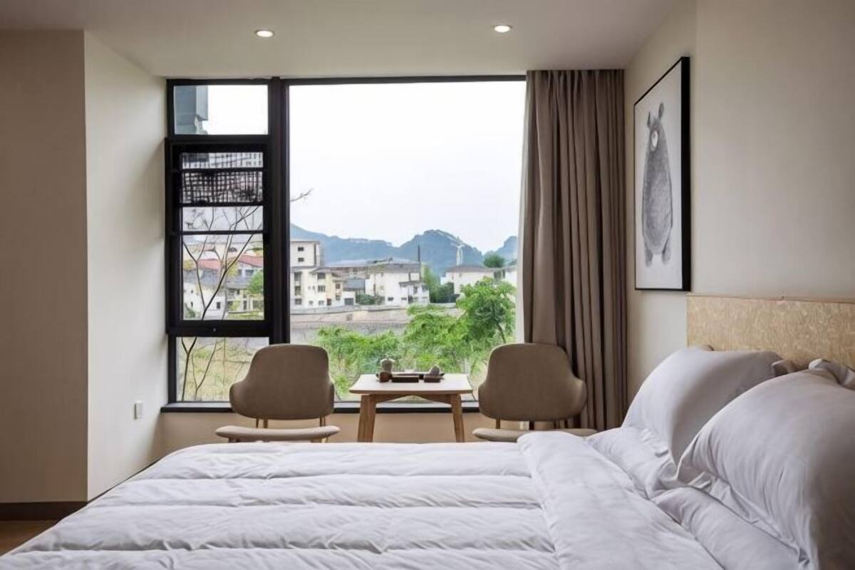 Ein Hotelzimmer mit einem großen Bett, zwei Stühlen, einem kleinen Tisch und einem Fenster mit Blick auf Gebäude und Berge.