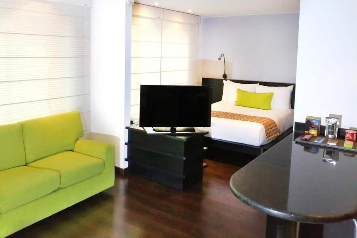 Quarto de hotel com um sofá verde, uma cama com roupa de cama branca e amarela, uma TV de tela plana em um suporte e uma mesa pequena com objetos sobre ela.